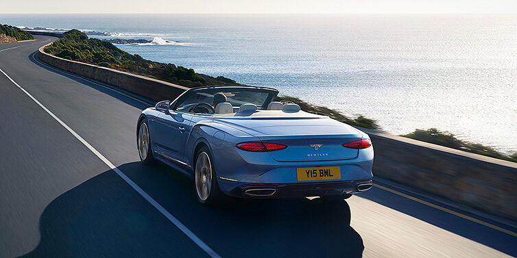 Bentley Tel Aviv New Bentley Continental GTC Azure convertible rear view in Light Windsor blue paint with 22 inch ten spoke wheels driving dynamically on an ocean road in the sunshine